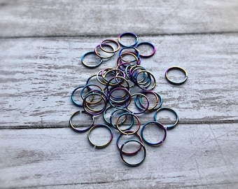 Puede incluir: Un conjunto de pequeños anillos metálicos iridiscentes, dispersos sobre una superficie de madera gris claro. Los anillos muestran un espectro de colores, incluyendo azules, verdes y morados, creando un efecto arcoíris. Probablemente sean joyas para el cuerpo, como piercings.