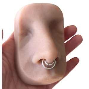 Puede incluir: Un modelo de nariz color carne con un anillo de tabique plateado. El modelo se sostiene en una mano sobre un fondo blanco. El anillo del tabique tiene un dise&ntilde;o de doble aro.