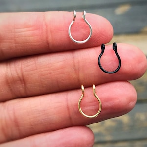 May include: Three nose rings in silver, black, and gold. The U-shaped rings are held between fingers, showcasing the different color options. The jewelry is designed to be worn on the nose.