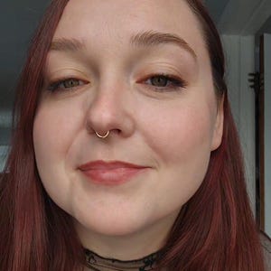 Puede incluir: Retrato en primer plano de una persona con piel clara, cabello rojo y un aro de nariz dorado. La persona est&aacute; sonriendo, con maquillaje visible, incluyendo l&aacute;piz labial y sombra de ojos. Se ve una blusa de encaje negro.