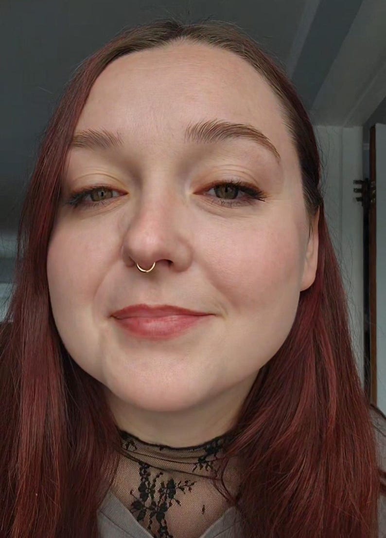 Puede incluir: Retrato en primer plano de una persona con piel clara, cabello rojo y un aro de nariz dorado. La persona est&aacute; sonriendo, con maquillaje visible, incluyendo l&aacute;piz labial y sombra de ojos. Se ve una blusa de encaje negro.