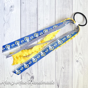 May include: A blue and yellow hair tie with a white and blue striped ribbon and a blue ribbon with a repeated pattern of a white ram's head. The hair tie is on a white wooden surface.