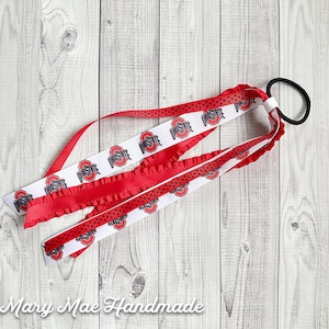 May include: A red, white, and black hair tie with Ohio State University logos on the ribbon. The hair tie is made of three different ribbons, one with a ruffled edge, one with black polka dots, and one with the Ohio State logo.