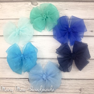 May include: Six different colored sheer ribbon bows arranged in a circle on a white wooden background. The bows are in shades of blue, teal, and light blue.