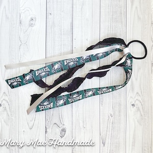 May include: A black hair tie with green and white ribbon featuring the Philadelphia Eagles logo. The ribbon is tied in a bow and the hair tie is on a white wooden background.