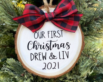 May include: A round wooden ornament with a red and black buffalo plaid bow. The ornament is decorated with white paint and the text "Our First Christmas Drew & Liv 2021".