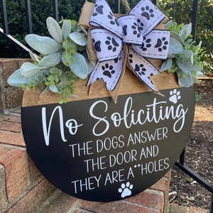 Op de afbeelding: Een ronde houten deurhanger met een zwarte en bruine achtergrond. Het bordje staat "No Soliciting The Dogs Answer The Door And They Are A**holes". Het bordje is versierd met een wit en zwart lint met poot afdrukken.