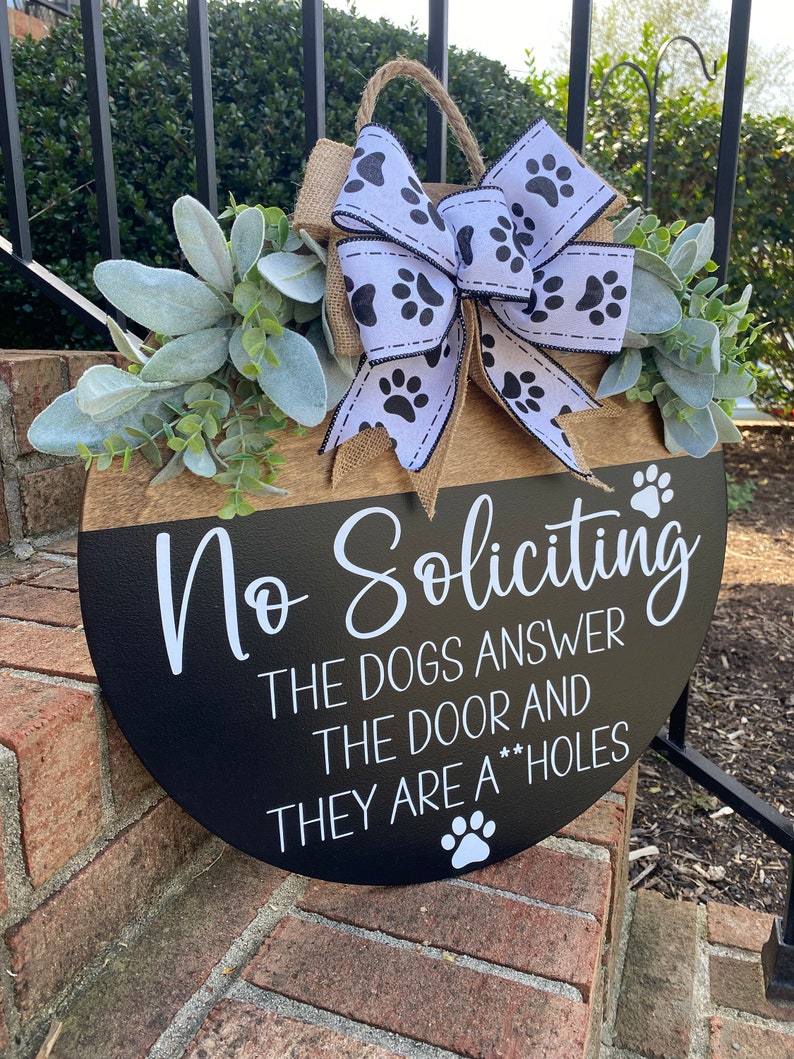 Op de afbeelding: Een ronde houten deurhanger met een zwarte en bruine achtergrond. Het bordje staat "No Soliciting The Dogs Answer The Door And They Are A**holes". Het bordje is versierd met een wit en zwart lint met poot afdrukken.