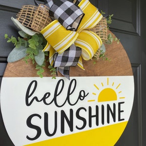 Może przedstawiać: Okrągły drewniany wieszak na drzwi z białym i ż&oacute;łtym tłem. Słowa "hello sunshine" są napisane czarnym kolorem z ż&oacute;łtym graficznym słońcem. Wieszak jest ozdobiony czarno-białą kratkowaną wstążką, ż&oacute;łto-białą wstążką w paski i jutowym kokardką.