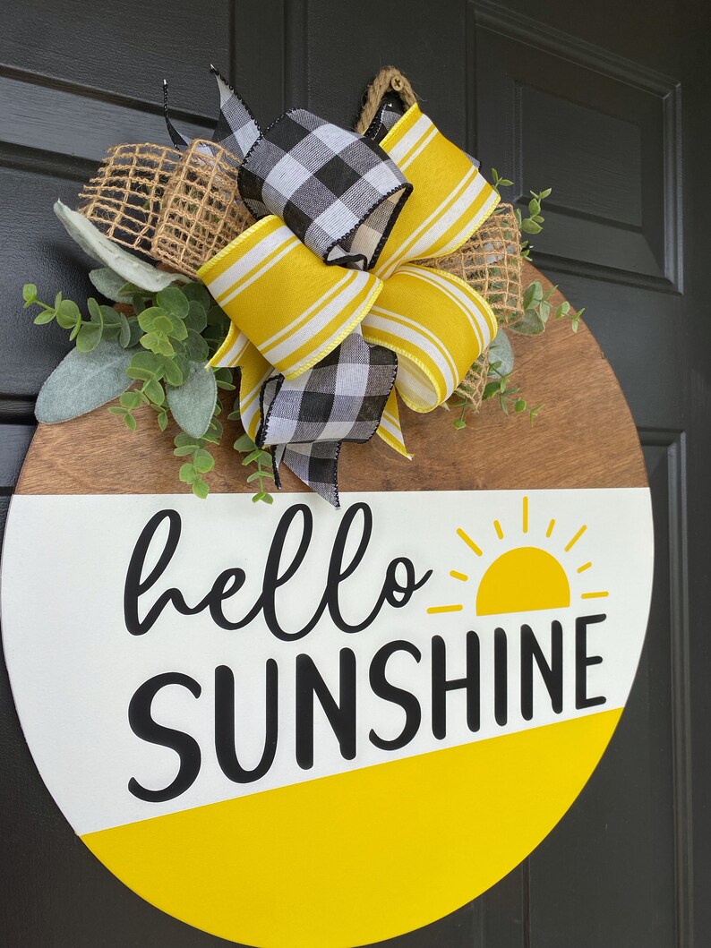 Może przedstawiać: Okrągły drewniany wieszak na drzwi z białym i ż&oacute;łtym tłem. Słowa "hello sunshine" są napisane czarnym kolorem z ż&oacute;łtym graficznym słońcem. Wieszak jest ozdobiony czarno-białą kratkowaną wstążką, ż&oacute;łto-białą wstążką w paski i jutowym kokardką.
