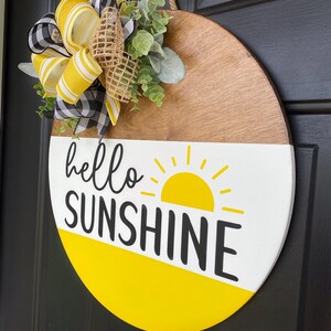 Może przedstawiać: Okrągły drewniany wieszak na drzwi z ż&oacute;łtym i białym tłem. Słowa "hello sunshine" są napisane czarnym kolorem z ż&oacute;łtym słonecznym wzorem. Wieszak jest ozdobiony ż&oacute;łto-czarną wstążką i zielenią.