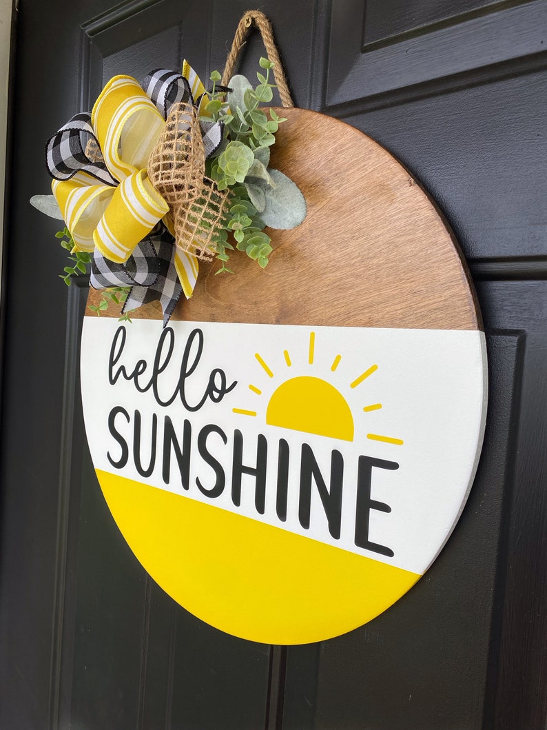 Może przedstawiać: Okrągły drewniany wieszak na drzwi z ż&oacute;łtym i białym tłem. Słowa "hello sunshine" są napisane czarnym kolorem z ż&oacute;łtym słonecznym wzorem. Wieszak jest ozdobiony ż&oacute;łto-czarną wstążką i zielenią.