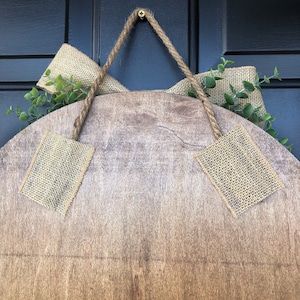 Op de afbeelding: Een rond houten bord met een jute lint en groen. Het bord is met een touw en een schroef aan een deur bevestigd.