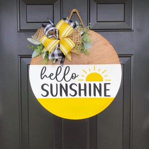 Może przedstawiać: Okrągły drewniany wieszak na drzwi z ż&oacute;łtym i białym wzorem słońca i tekstem "hello sunshine". Wieszak jest ozdobiony ż&oacute;łtą i czarną kratkowaną kokardką i zielenią.