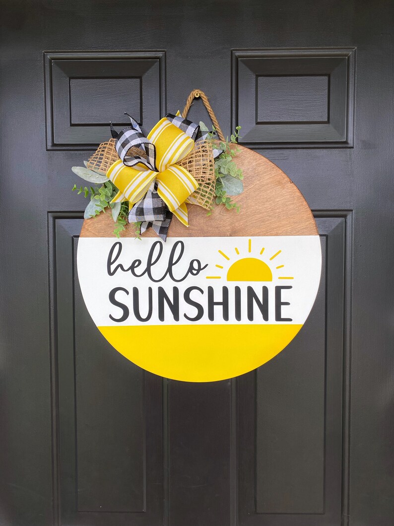 Może przedstawiać: Okrągły drewniany wieszak na drzwi z ż&oacute;łtym i białym wzorem słońca i tekstem "hello sunshine". Wieszak jest ozdobiony ż&oacute;łtą i czarną kratkowaną kokardką i zielenią.