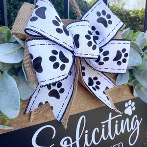 Op de afbeelding: Een houten bord met een zwarte achtergrond en witte tekst die "Soliciting Dogs Answer" luidt. Het bord is versierd met een jute strik met een wit lint met zwarte poot afdrukken.