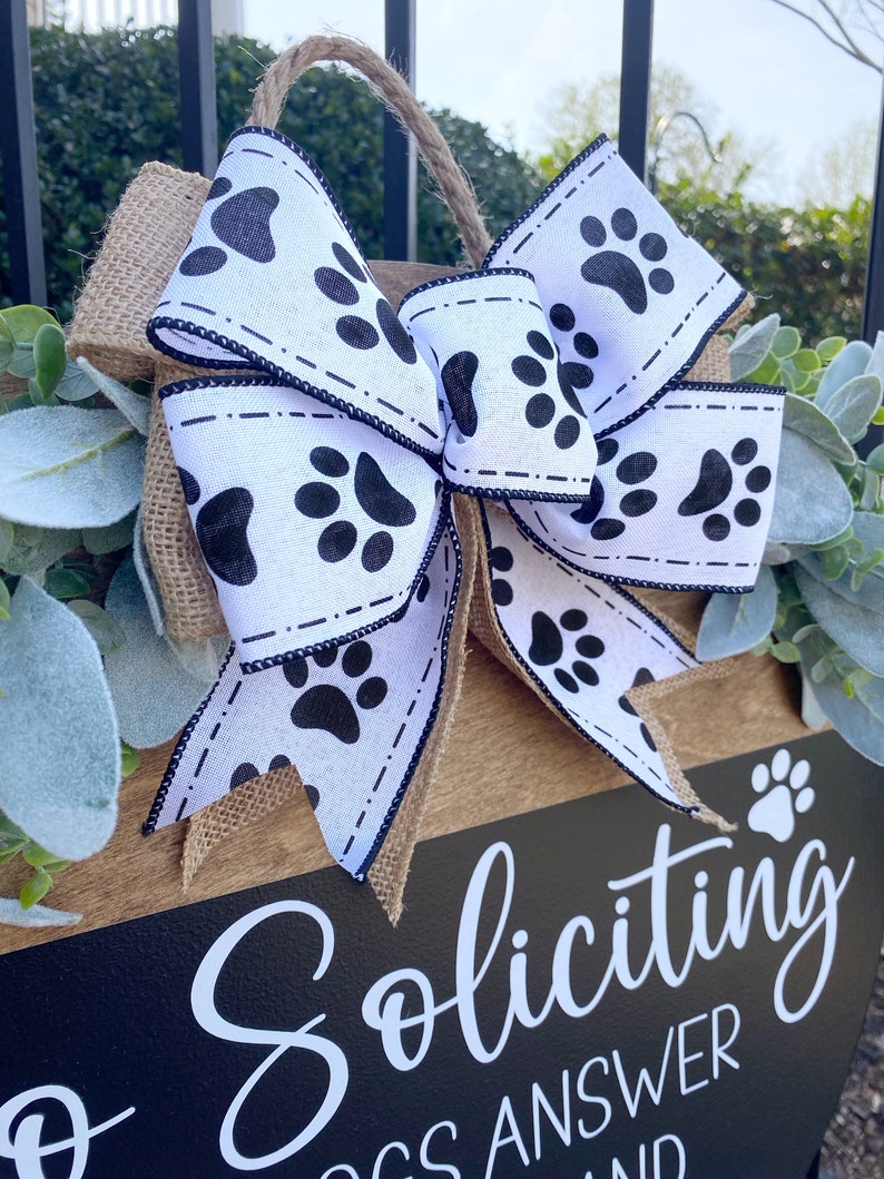 Op de afbeelding: Een houten bord met een zwarte achtergrond en witte tekst die "Soliciting Dogs Answer" luidt. Het bord is versierd met een jute strik met een wit lint met zwarte poot afdrukken.