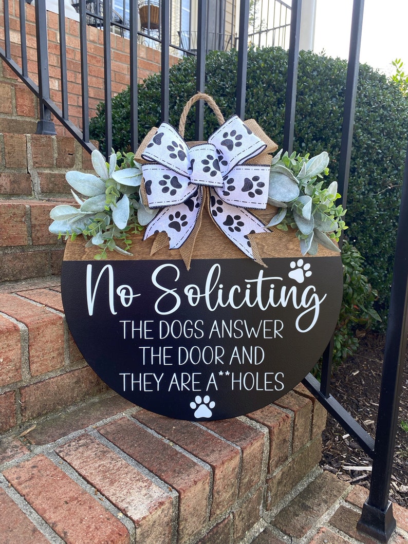 Op de afbeelding: Een rond houten deurschild met een zwarte achtergrond en witte tekst die "No Soliciting The Dogs Answer The Door And They Are A**holes" luidt. Het bord is versierd met een jute strik met witte poot afdrukken en een groene krans van bladeren.