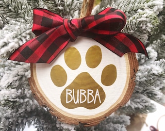 May include: A wooden Christmas ornament with a gold paw print and the name "Bubba" written in white. The ornament is decorated with a red and black plaid bow.