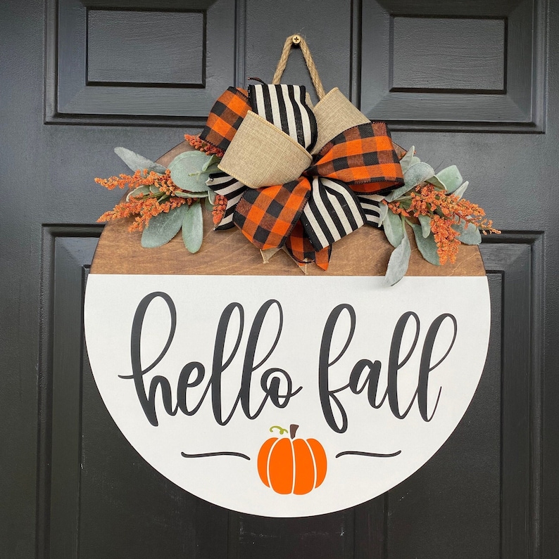 May include: A round wooden door hanger with a white background. The door hanger has a brown wooden top half and a white bottom half. The white bottom half has the words 'hello fall' written in black cursive script. There is a small orange pumpkin with a green stem below the words. The top half of the door hanger has a brown burlap bow with black and orange checkered ribbon and green leaves.