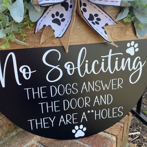 Op de afbeelding: Een zwart-wit bord met de tekst "No Soliciting The Dogs Answer The Door And They Are A**holes" met een pootprint aan de onderkant. Het bord is versierd met een zwart-witte strik met pootprintjes.