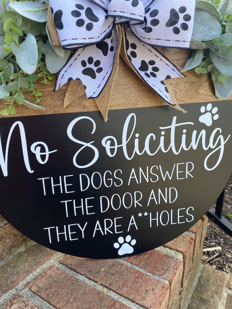 Op de afbeelding: Een zwart-wit bord met de tekst "No Soliciting The Dogs Answer The Door And They Are A**holes" met een pootprint aan de onderkant. Het bord is versierd met een zwart-witte strik met pootprintjes.