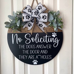 Op de afbeelding: Een ronde houten deurhanger met een zwarte achtergrond en witte tekst die "No Soliciting The Dogs Answer The Door And They Are A**holes" luidt. De hanger is versierd met een wit lint met zwarte poot afdrukken en een groene krans van bladeren.
