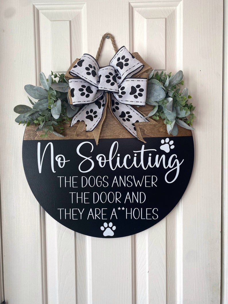 Op de afbeelding: Een ronde houten deurhanger met een zwarte achtergrond en witte tekst die "No Soliciting The Dogs Answer The Door And They Are A**holes" luidt. De hanger is versierd met een wit lint met zwarte poot afdrukken en een groene krans van bladeren.