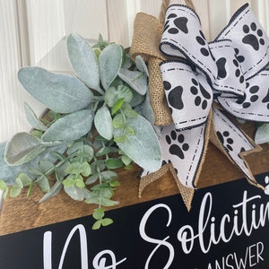 Op de afbeelding: Een zwart-wit deur bord met de tekst "No Soliciting" en "The dogs answer the door and bark". Het bord is versierd met een jute strik met een wit lint met zwarte poot afdrukken. Het bord is ook versierd met een takje groen en grijs gebladerte.