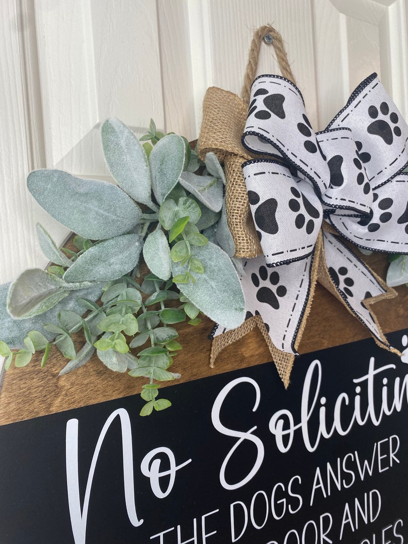 Op de afbeelding: Een zwart-wit deur bord met de tekst "No Soliciting" en "The dogs answer the door and bark". Het bord is versierd met een jute strik met een wit lint met zwarte poot afdrukken. Het bord is ook versierd met een takje groen en grijs gebladerte.