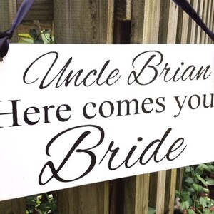 May include: White sign with black text that reads "Uncle Brian Here comes your Bride". The sign is hanging from a wooden fence with black ribbon.