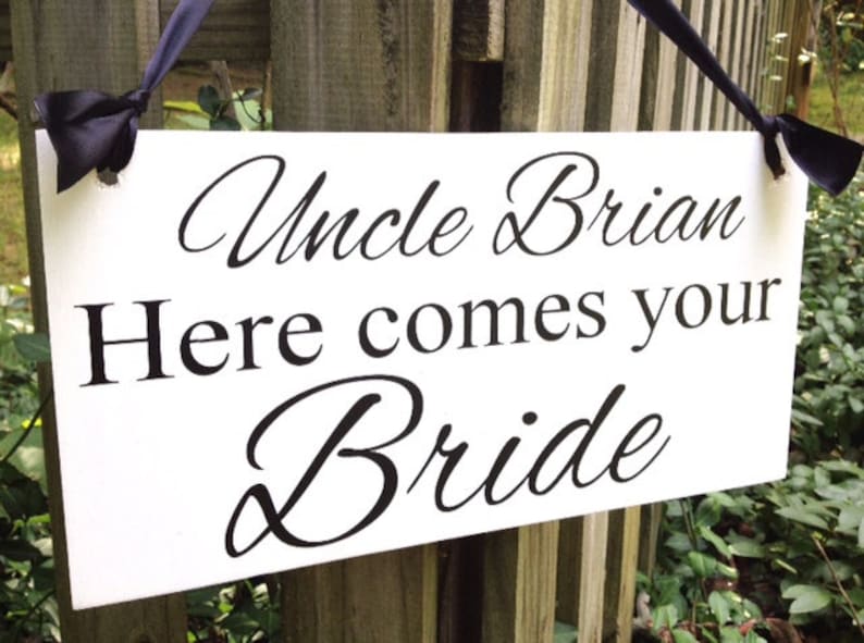 May include: White sign with black text that reads "Uncle Brian Here comes your Bride". The sign is hanging from a wooden fence with black ribbon.
