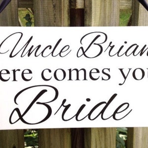 May include: White sign with black text that reads "Uncle Brian Here comes your Bride". The sign is hanging from a black ribbon.