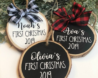 May include: Three round wood ornaments with black and white checkered ribbon, red and black checkered ribbon, and a black and white ribbon. The ornaments are painted white and black and have the text "Noah's First Christmas 2019", "Ava's First Christmas 2019", and "Olivia's First Christmas 2019".