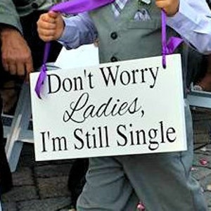 May include: A young boy in a gray suit and a purple ribbon holds a white sign that says "Don't Worry Ladies, I'm Still Single".