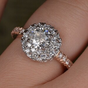 May include: A rose gold engagement ring with a large, round, clear diamond center stone surrounded by a halo of smaller diamonds. The band is also adorned with small diamonds, creating a sparkling effect. The ring is on a person's finger.