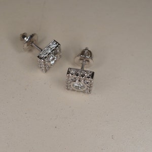 May include: A pair of square-shaped stud earrings. Each earring features a central, round, clear gemstone surrounded by smaller, clear gemstones. The earrings are set in a silver-colored metal and are displayed on a light-colored surface.