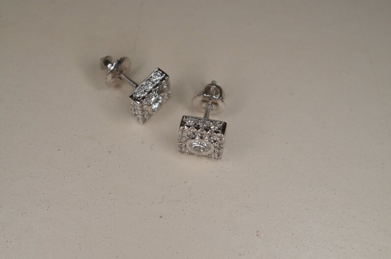 May include: A pair of square-shaped stud earrings. Each earring features a central, round, clear gemstone surrounded by smaller, clear gemstones. The earrings are set in a silver-colored metal and are displayed on a light-colored surface.
