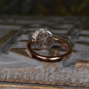 May include: Rose gold ring with a round, diamond-encrusted halo setting. The band features a delicate, twisted design. The ring is set against a textured, white and gold surface, showcasing its intricate details and elegant design.