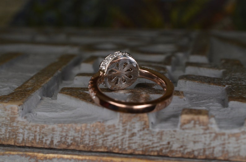 May include: Rose gold ring with a round, diamond-encrusted halo setting. The band features a delicate, twisted design. The ring is set against a textured, white and gold surface, showcasing its intricate details and elegant design.