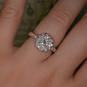 May include: A rose gold engagement ring with a large, round, diamond halo. The center stone is surrounded by smaller diamonds, and the band is also set with diamonds. The ring is on a person's finger.