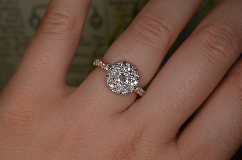 May include: A rose gold engagement ring with a large, round, diamond halo. The center stone is surrounded by smaller diamonds, and the band is also set with diamonds. The ring is on a person's finger.