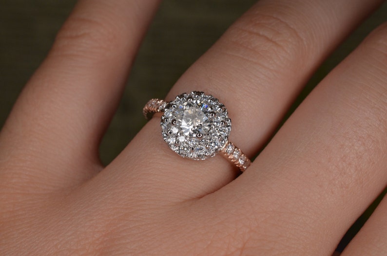 May include: A rose gold engagement ring with a large, round, clear diamond center stone surrounded by a halo of smaller diamonds. The band is also adorned with small diamonds, creating a sparkling effect. The ring is on a person's finger.