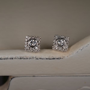 May include: A pair of square-shaped stud earrings. Each earring features a central, round, clear gemstone surrounded by smaller, clear gemstones. The earrings are set in a silver-colored metal and rest on a white surface.