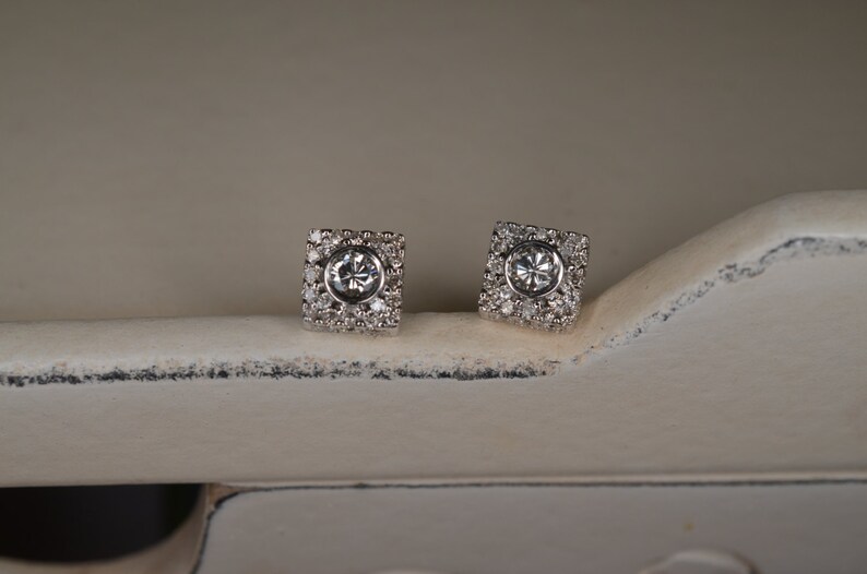 May include: A pair of square-shaped stud earrings. Each earring features a central, round, clear gemstone surrounded by smaller, clear gemstones. The earrings are set in a silver-colored metal and rest on a white surface.