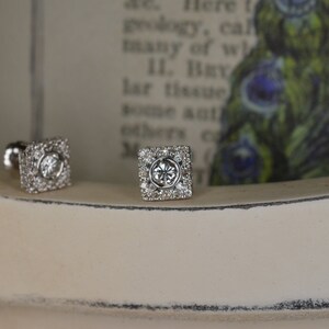 May include: A pair of square-shaped cufflinks with a central clear stone surrounded by smaller, clear stones. The cufflinks are set on a white surface, with a blurred background featuring text and a peacock feather design.