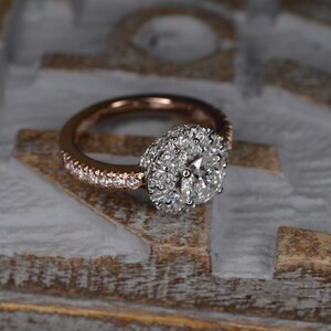 May include: Rose gold engagement ring with a halo of small diamonds surrounding a larger central diamond. The band is also encrusted with small diamonds. The ring is set against a textured, light-colored background.