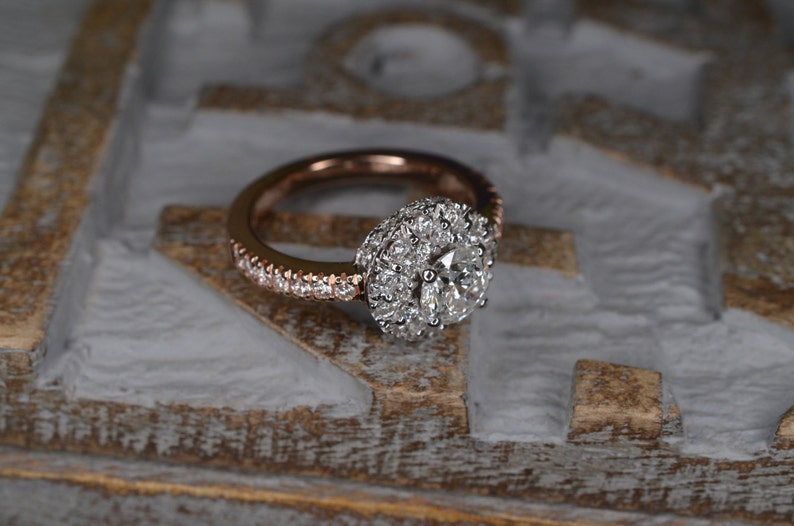 May include: Rose gold engagement ring with a halo of small diamonds surrounding a larger central diamond. The band is also encrusted with small diamonds. The ring is set against a textured, light-colored background.