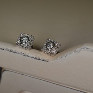 May include: A pair of square-shaped stud earrings. Each earring features a central, round, clear gemstone surrounded by smaller, clear gemstones. The earrings are set in a silver-colored metal and rest on a distressed, off-white surface.