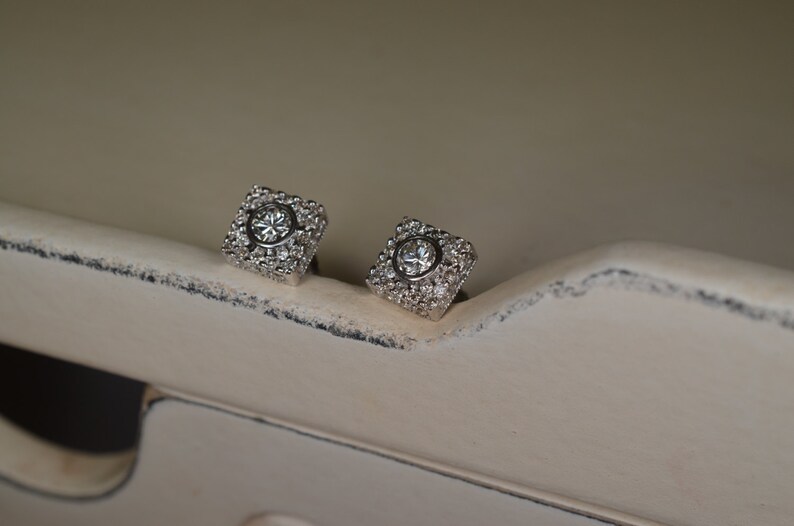 May include: A pair of square-shaped stud earrings. Each earring features a central, round, clear gemstone surrounded by smaller, clear gemstones. The earrings are set in a silver-colored metal and rest on a distressed, off-white surface.